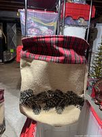 Front view of one of the beige fabric Christmas baskets with pine cone and pine branch design, featuring red plaid trim on the folded top edge.