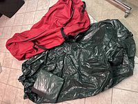 Three large Christmas tree or garland storage bags laid out on the floor, two green plastic-free bags and one red canvas-like bag.