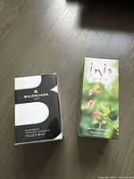 Photo showing both perfume boxes side by side on floor: black and white Balenciaga and green nature-themed Inis Arose.