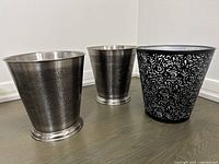 Three metal waste baskets arranged upright on floor showing front and side details, two silver with engraved floral patterns and one black with cutout floral designs.