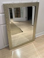 Front view of rectangular beveled mirror with textured champagne gold square-patterned frame leaning against white door.