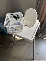 White IKEA high chair with plastic seat, tray, and metal legs seen from an angle. A white basket on the tray contains corner guards in packaging.