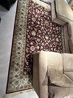 Full view of large maroon rug with beige floral border under sectional sofa.