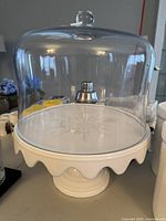 White ceramic cake stand with scalloped edge and clear glass dome lid with knob on top, shown side view.