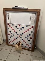 Large framed magnetic Scrabble board showing playing grid and frame, with a beige drawstring bag placed at the base containing tiles.