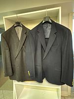 Two men's wool suit jackets hanging side by side on hangers, showing front view and full length for color and pattern visibility.
