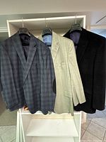 Three men's wool suit jackets hanging on hangers showing colors and patterns including grey plaid, beige, and black.