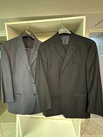 Two men's wool suits hanging on hangers, one grey with check pattern, the other black with pinstripes.