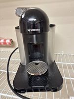 Front view of black Nespresso Vertuo machine with silver water tank and power cord visible