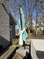 Full view of the folded large teal square umbrella attached to brown metal pole and heavy base, located outdoors beside patio furniture and house exterior.