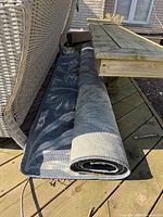 Rolled up blue and teal tropical floral outdoor rug beside outdoor furniture on wooden deck.