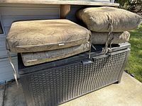 Stack of four beige outdoor pillow cushions with leafy pattern fabric on top of brown Rubbermaid plastic storage bench outside on concrete.