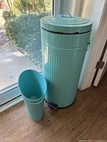 View of the large teal metal garbage bin with lid, foot pedal, and the smaller teal plastic bin next to it by a glass door.
