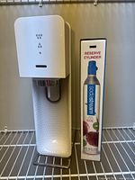 Frontal view of white SodaStream soda maker with textured panel and reserve cylinder box alongside.