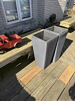 Two tall dark gray plastic wicker style planters placed side by side on wooden deck.