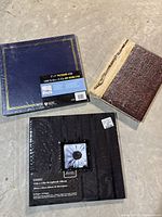 Three photo albums: one blue and gold 12x12 album, one brown and cream vintage style album, and one black 12x12 scrapbook album with flower photo window.