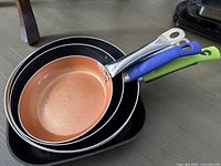 Stacked view of three non-stick frying pans in copper and black interiors with metal and colored handles