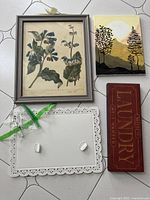 Lot contents photographed on floor with white tile background. Items laid out: framed floral botanist print, painted forest scene canvas, Laundry sign, and white metal bulletin board with magnets.