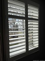 Two white shutter panels with horizontal louvers partially opened, displaying outdoor view through window.