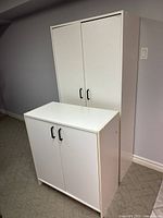 Photo of two white storage cabinets, one smaller and one taller, both with double doors and black handles, placed on carpet.