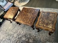 Three wooden tables on a rug: one square coffee table in center and two side tables flanking it, showing parquet pattern tops and carved legs.