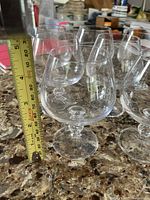 Seven clear brandy snifter glasses arranged on a countertop with a measuring tape showing roughly 4.5 inch height.