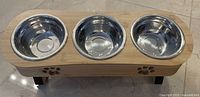 Wooden cat bowl holder with three stainless steel bowls inserted, showing the wood grain, black legs, and bowl details.