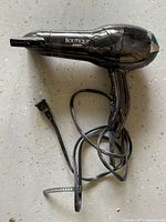 Black Conair Boutique hair dryer with concentrator nozzle and attached power cord on a speckled grey floor.
