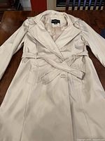 Front of cream coloured 3/4 length jacket laid flat on wooden surface, shows collar, double-breasted buttons, belt, and pockets.