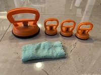 Four orange plastic dent pullers in different sizes and a light blue microfiber cloth placed on a floor surface.