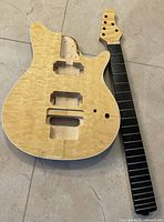 Photo showing the guitar body and neck placed side by side on a flat surface. Body is natural quilted maple top with cutouts, neck dyed dark rosewood with maple headstock and ebony fretboard without frets.