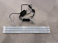 Four slim 12-inch LED light bars laid out on a carpeted floor, connected to a black power adapter with switch through wires.