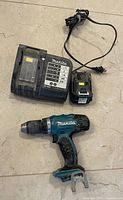 Photo shows Makita cordless drill, battery pack, and battery charger on a floor background.