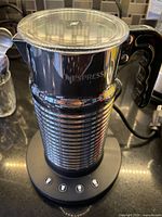 Front angled view of the Nespresso Aeroccino 4 milk frother placed on its base showing ribbed stainless steel body, clear lid, black handle, and control buttons on base.