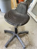 Black pottery wheel saddle seat with five-legged base on concrete floor, showing minor paint and splatter marks.