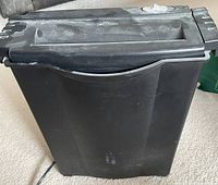 Photo showing a black six sheet paper shredder, dusty on top and front surfaces with visible power cord, placed on carpet floor.
