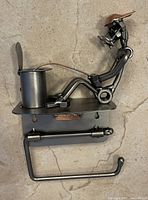 Front view of black metal steampunk-style toilet paper holder featuring a figure sitting with a laptop, mounted on a rectangular base with a toilet paper rod below.