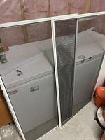 Pair of window screens leaning on a white freezer in an unfinished basement or storage area, showing the full frames and mesh material intact with no visible tears.