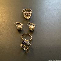 Photo of three pieces of jewelry: a prism cube ring with a color-changing faceted stone, a heart-shaped faceted ring, and a pair of silver-tone spherical drop earrings.