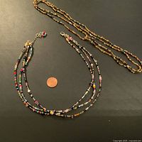 Two vintage glass bead necklaces displayed on black background with a penny for scale. The necklaces are a double-strand colorful bead necklace with clasp and a long amber-toned bead necklace.