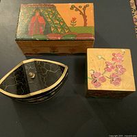 Three vintage hand-painted trinket or jewelry boxes shown closed: rectangular wooden box with traditional figure and floral painting, square wooden box with pink blossoms on lid, and black and white leaf-shaped box.