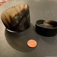 Photo of two horn cuff bangles lying flat next to a penny for size reference, showing natural horn coloration and polished finish.