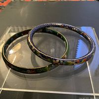 Two vintage cloisonné bangles, one dark blue and one green, displayed on a clear stand.