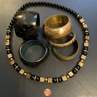 Photo showing set of five bracelets and one beaded necklace laid out on a dark surface with a penny for scale. Bracelets vary in material and design with black, brass, and green colors, and necklace has brass and black beads.