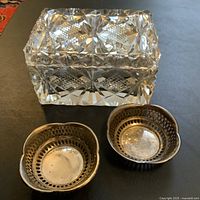 Photo showing one cut crystal rectangular jewelry box with lid and two smaller round silver-tone trinket dishes with pierced lattice sides, all on a dark surface.