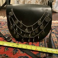 Front view of vintage black leather crossbody bag with three chains of metal coin charms draped on front flap; adjustable leather strap seen.