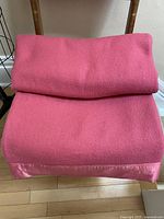 Folded warm pink 100% virgin wool blanket with satin binding displayed on a wooden chair and floor.