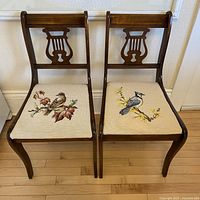Front photo showing two vintage wooden chairs side by side with carved lyre-style backrests and hand stitched needlepoint bird seat covers.