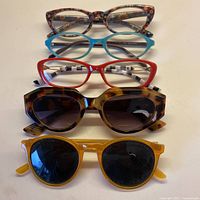 Five pairs of glasses displayed together: three pairs of reading glasses on top and two pairs of sunglasses below, showing various frame styles and colors.