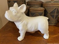 Side profile of white ceramic Frenchie piggy bank on wooden surface with decorative containers in background.